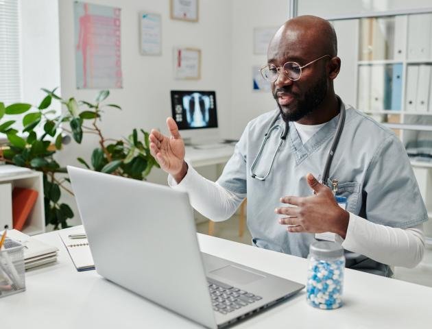 Young confident African American online doctor in uniform sitting by desk in front of laptop in clinics and giving medical advice to patient